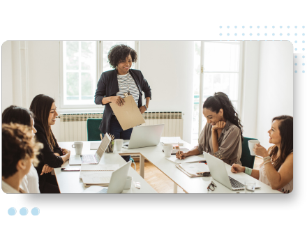 A diverse team in a bright office brainstorms around laptops. A woman stands confidently, holding a folder, engaging six colleagues seated at a table. Highlighting tailored pre-employment assessments for effective talent acquisition.